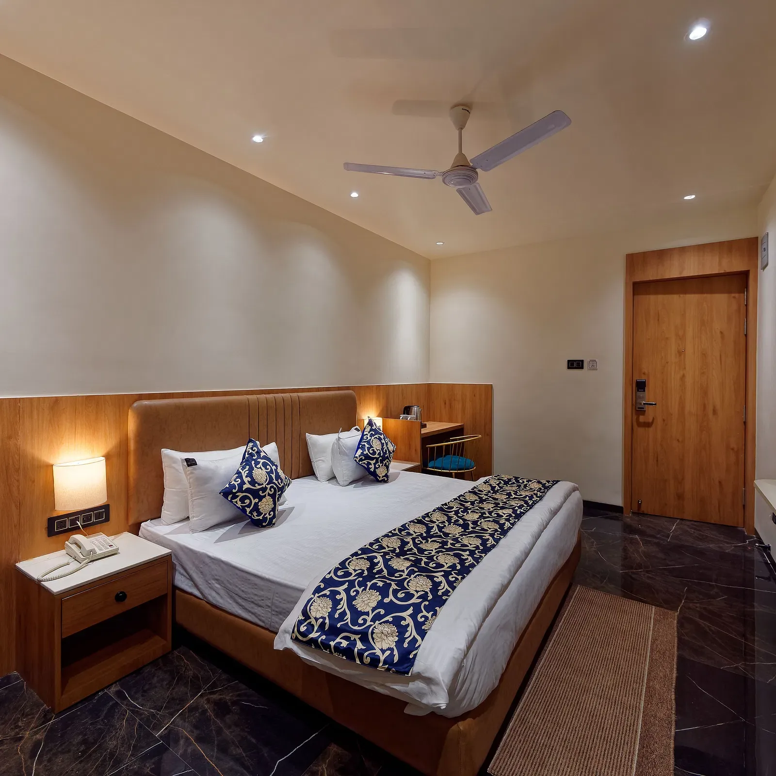 Image of a studio room with a bed with a table on the side of the bed and a television in front Image of a bed with table and chair on the side of the table and a fan on te ceiling at Ankit Hotel, Jabalpur